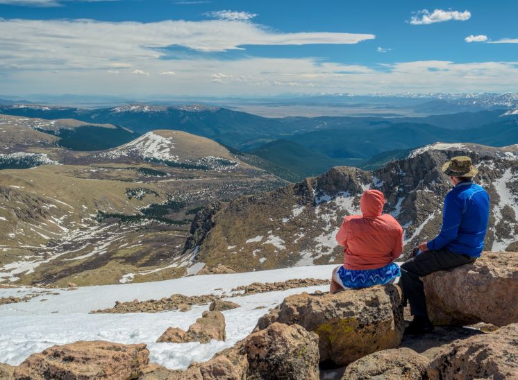 A couple sitting on a rock admiring the view of the Rocky Mountains from the snow covered sumit of Mount Evans